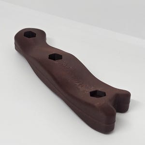 May include: A dark brown wrench-like tool with three hexagonal openings. The tool has a curved handle and a forked end. The object is made of a solid material and is designed for tightening or loosening nuts and bolts.
