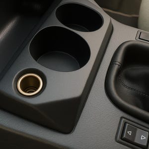 Puede incluir: Primer plano del interior de un coche, que muestra una consola negra con dos portavasos y un encendedor de cigarrillos. El encendedor tiene un interior dorado. También se ven una palanca de cambios y un panel de control.