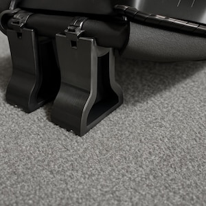 Puede incluir: Primer plano de un objeto de metal negro con acabado mate. El objeto tiene una serie de soportes rectangulares y una sección superior curvada. Los soportes están conectados por bisagras. El fondo es una superficie gris texturizada.