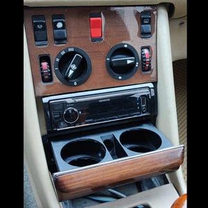 May include: Car interior with a wood-grain dashboard. Features include a Kenwood stereo, various control knobs, and a retractable cup holder. The dashboard has black and red buttons and switches.
