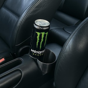 Puede incluir: Una lata negra y verde de Monster Energy en un portavasos de coche. La lata tiene una tapa plateada y el logotipo de Monster en verde. El portavasos es de acero inoxidable y está situado en un interior de coche de cuero negro.