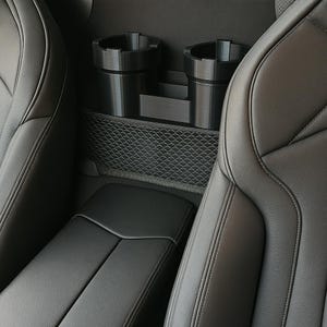 Pode incluir: Um porta-copos e organizador de armazenamento preto para carro instalado entre dois assentos de couro cinza. O organizador tem dois porta-copos e um pequeno compartimento de armazenamento. Há um bolso de malha abaixo.