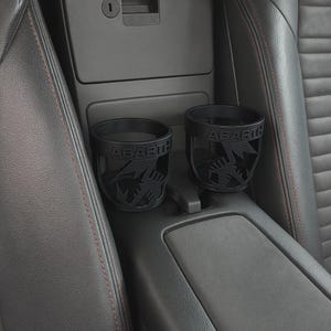 May include: Two black car cup holders with the word "ABARTH" and a scorpion logo. The cup holders are placed in a car's centre console, surrounded by black leather seats with red stitching.