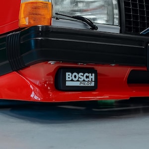 Puede incluir: Primer plano de un parachoques rojo con un detalle negro. Un emblema rectangular negro de Bosch Pilot está centrado en el parachoques. Se ven el faro y el intermitente del coche.