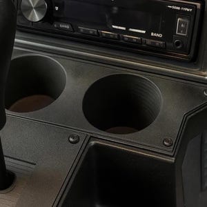 Puede incluir: Primer plano del interior negro de un coche con una radio, dos portavasos y una palanca de cambios. La radio tiene una pantalla digital y botones. Los portavasos son circulares. La palanca de cambios es negra.