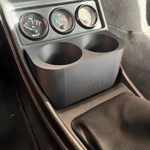 Peut inclure: Un porte-gobelets noir avec deux emplacements pour gobelets et le mot "GIII" imprimé sur le devant. Le porte-gobelets est installé dans la console centrale d'une voiture, à côté de trois jauges rondes.