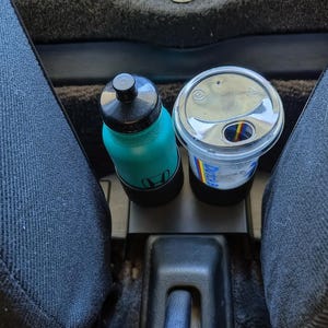 May include: A teal water bottle with a black cap and a white Honda logo sits next to a clear plastic cup with a lid. The cup has a colorful logo and is in a car's center console, between two dark blue seats.