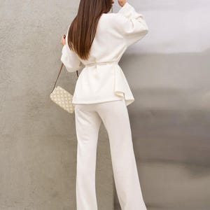 May include: A cream-colored two-piece outfit consisting of a long-sleeved top with a belted waist and wide-leg pants. The outfit is paired with a small, patterned handbag. The model is facing away from the camera.