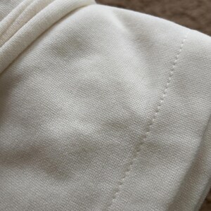 May include: Close-up of a cream-colored fabric, possibly a garment, with a visible seam and textured surface. The fabric appears soft and slightly wrinkled, suggesting a comfortable material. The background is a blurred brown.