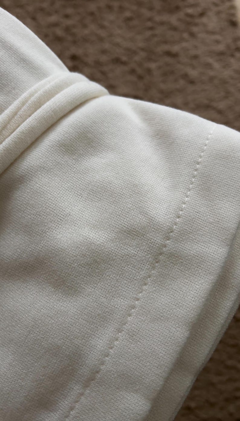 May include: Close-up of a cream-colored fabric, possibly a garment, with a visible seam and textured surface. The fabric appears soft and slightly wrinkled, suggesting a comfortable material. The background is a blurred brown.