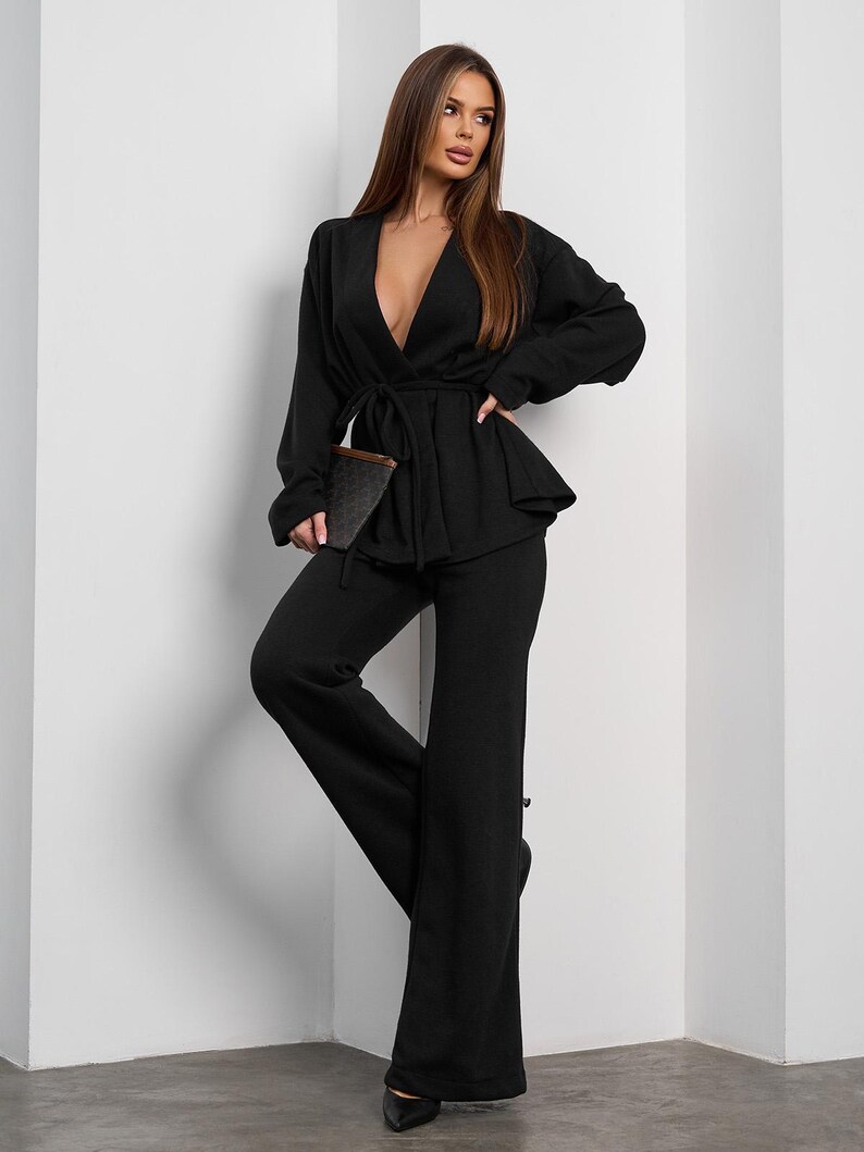 May include: A black two-piece outfit featuring a wrap top with a tie waist and wide-leg pants. The outfit includes a matching clutch and is paired with black heels. The ensemble is a monochromatic, elegant style.