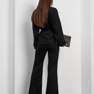 May include: A black two-piece outfit featuring a long-sleeved top with a waist tie and flared pants. The outfit is paired with a dark clutch. The model is facing away from the camera, showcasing the back of the outfit.
