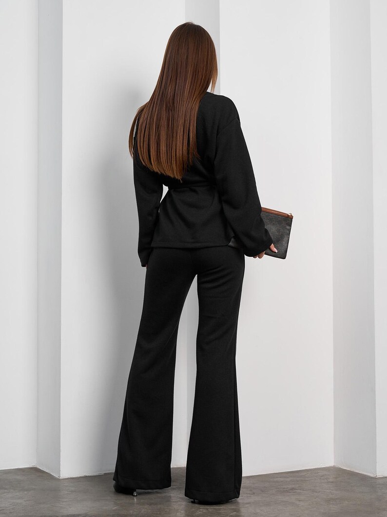 May include: A black two-piece outfit featuring a long-sleeved top with a waist tie and flared pants. The outfit is paired with a dark clutch. The model is facing away from the camera, showcasing the back of the outfit.