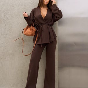 May include: A brown two-piece outfit consisting of a wrap top with a waist tie and wide-leg pants. A small, tan handbag with a shoulder strap is held in the hand. The outfit is made of a soft, textured fabric.