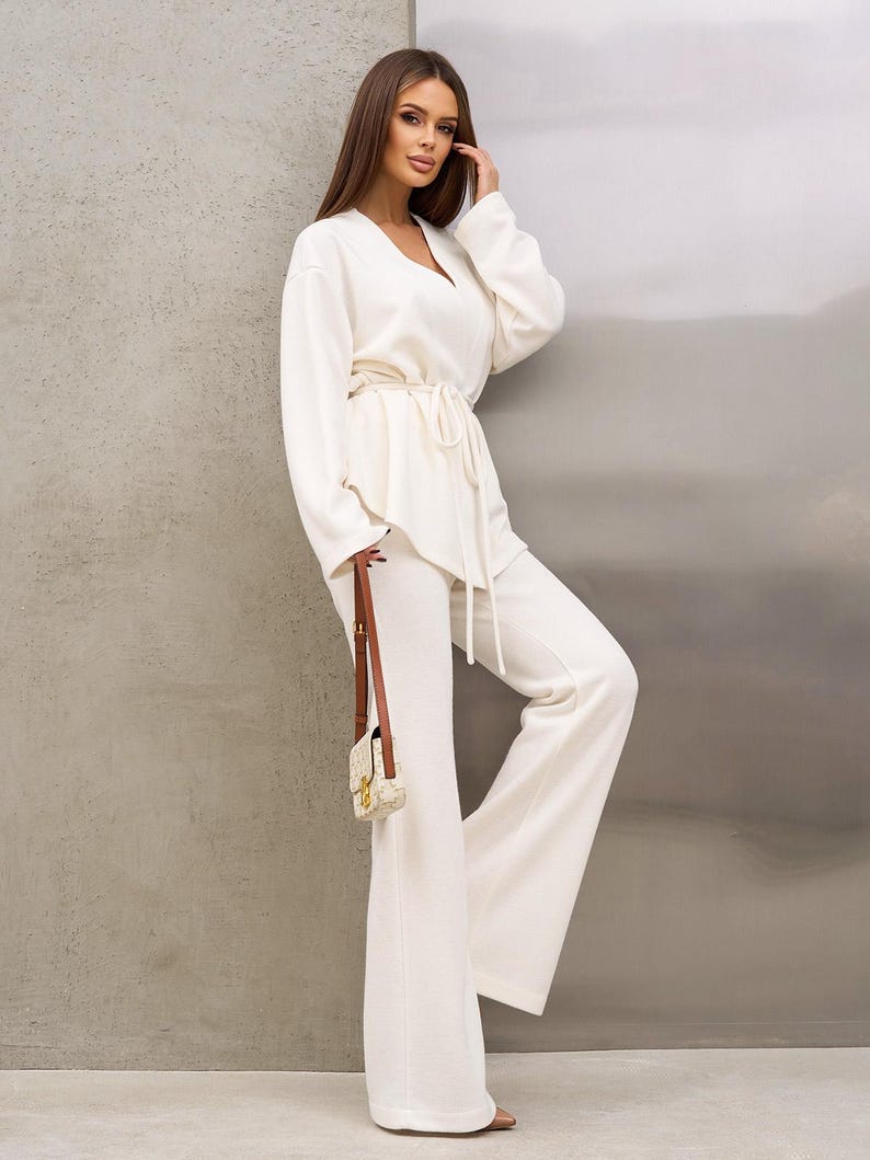 May include: A cream-colored, two-piece outfit featuring a long-sleeved top with a V-neck and a drawstring waist, paired with wide-leg pants. A small, patterned purse with a brown strap is held. The outfit is made of a soft, textured fabric.