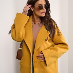 Puede incluir: Un abrigo amarillo mostaza con solapas grandes y dos botones. El abrigo tiene mangas largas y un corte entallado. Un bolso marrón se lleva sobre el hombro. La persona lleva gafas de sol y una camiseta marrón.