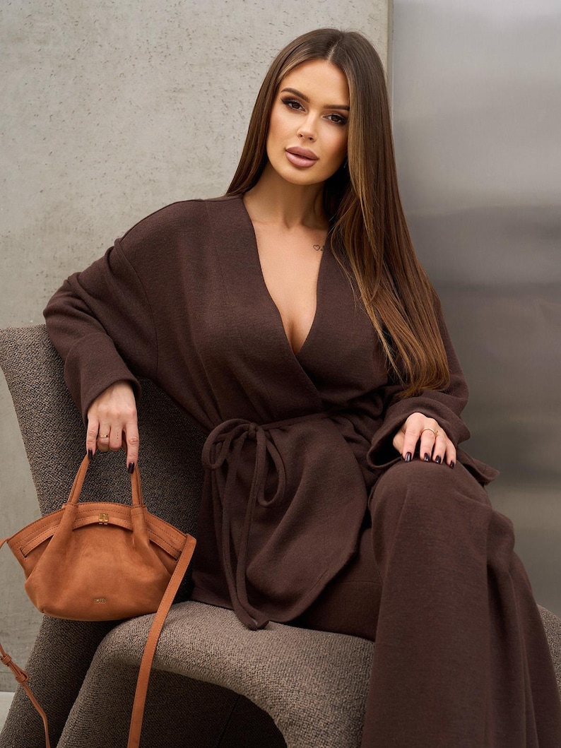 May include: A woman wearing a brown wrap-style top and matching wide-leg pants. She is seated on a gray chair and holding a tan handbag with a shoulder strap. The outfit is a solid color, and the handbag has a curved shape.