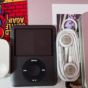 May include: A black iPod with a white docking station, white earbuds, and a silver case. The iPod has a screen and a circular control pad with a menu button. The earbuds are coiled. A colorful poster is in the background.