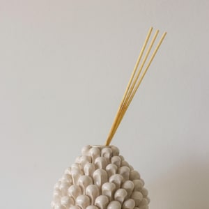 May include: A cream-colored ceramic essential oil diffuser with a unique, textured design. The diffuser has a cone-like shape, covered in small, rounded, droplet-like shapes. Three light-colored reeds are inserted into the top, ready to diffuse fragrance.