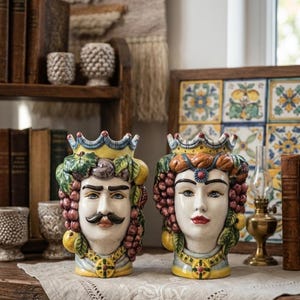 Può includere: Due vasi in ceramica a forma di testa raffiguranti una figura maschile e una femminile, ciascuna con una corona gialla. Le figure sono decorate con uva, limoni e foglie verdi scolpite. Entrambi i vasi presentano tratti del viso dipinti.
