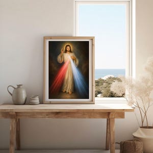 K&ouml;nnte beinhalten: Gerahmtes religi&ouml;ses Kunstwerk, das eine Figur in wei&szlig;en Gew&auml;ndern mit goldenem Heiligenschein zeigt, die rotes und blaues Licht ausstrahlt. Das Kunstwerk wird auf einem Holztisch neben einem Fenster mit Blick auf das Meer ausgestellt.