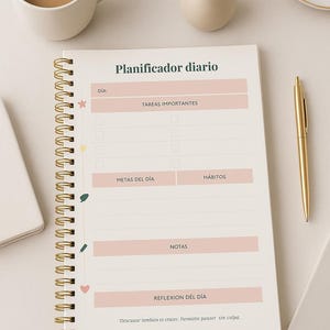 May include: A spiral-bound daily planner with gold accents, featuring sections for tasks, goals, and notes. The planner includes text in Spanish, a gold pen, a cup of coffee, and a small vase with dried flowers.
