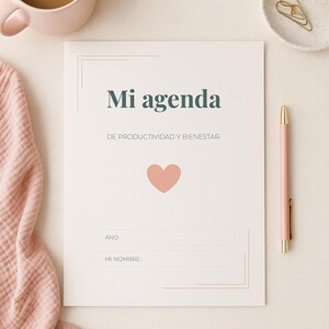May include: A planner with the text "Mi agenda" and a heart graphic. A pink pen, a cup of coffee, a rose, and a small dish with a gold paperclip are also visible. The planner includes sections for the year and name.