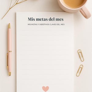 May include: A flat lay image featuring a pink mug with coffee, a notepad with the text "Mis metas del mes", a pink pen, dried flowers, and gold paper clips. The notepad has a pink heart at the bottom.