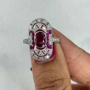 May include: An Art Deco-style ring featuring a large oval ruby center stone, surrounded by smaller rubies and clear accent stones. The ring has a silver-colored band and a rectangular face with geometric details. The design is symmetrical and elegant.