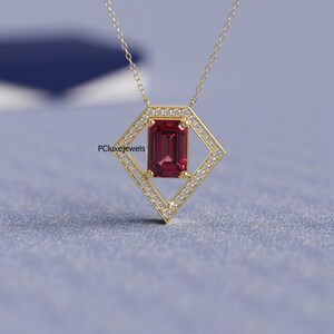May include: A gold necklace featuring a rectangular, deep red gemstone pendant. The pendant is set within a diamond-shaped frame, encrusted with small, clear gemstones. The necklace has a delicate chain and is displayed against a blue background. The text "PCluxejewels" is visible.