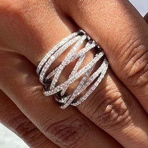 Puede incluir: Un anillo plateado con un diseño tejido, incrustado con numerosas piedras claras y brillantes. El intrincado patrón del anillo presenta múltiples bandas que se cruzan, creando una apariencia texturizada y llamativa. El anillo se muestra en un dedo.