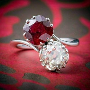 May include: A silver ring featuring a large, faceted, deep red ruby and a clear, sparkling diamond. The ring's setting is intricate, with the gemstones prominently displayed against a red background.