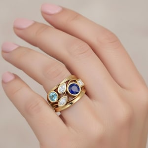 May include: A gold ring featuring a cluster of gemstones. The ring has a large, round, deep blue sapphire, a light blue gemstone, and several clear diamonds. The ring is on a hand with pink nail polish.
