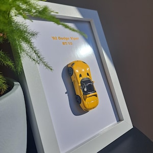 Puede incluir: Un coche de modelo a escala amarillo '92 Dodge Viper RT/10 enmarcado. El coche se muestra en un marco blanco sobre un fondo blanco. El texto "'92 Dodge Viper RT/10" está impreso en amarillo. El coche tiene ruedas negras e interior negro.