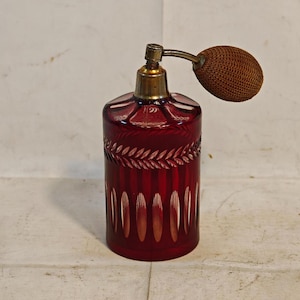Può includere: Un flacone di profumo in vetro rosso intenso con corpo cilindrico, motivi a taglio verticale e un motivo a foglie orizzontale. Il flacone è sormontato da un collare metallico e da una pompetta in tessuto marrone fissata da un beccuccio curvo.