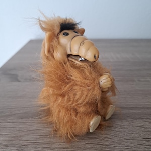 Op de afbeelding: Een vintage ALF-beeldje. De figuur heeft een bruin gezicht met zwarte ogen en een grote neus. Het lichaam is bedekt met ruige, bruine vacht. De figuur zit met gevouwen handen en uitgestrekte voeten.