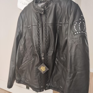 Puede incluir: Chaqueta de cuero negro con costuras decorativas de cadena a lo largo de la cremallera, el cuello y los puños. Cuenta con un emblema de corona en la manga izquierda y pequeños tachuelas plateadas. Una etiqueta con un logotipo de corona cuelga de la cremallera.