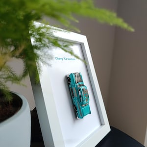 Peut inclure: Un cadre blanc avec une voiture jouet bleu turquoise à l'intérieur. La voiture est une Chevy '53 Custom. Le cadre est sur une surface noire.