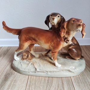 Puede incluir: Figura de cerámica con dos perros en tonos marrón y beige. Los perros están posados sobre una base clara con aspecto texturizado. La figura tiene un acabado brillante y se encuentra sobre un fondo neutro.