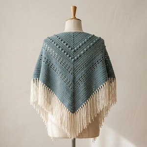 May include: A dusty blue triangular crochet shawl featuring a textured pattern of raised bobble stitches and openwork lace sections. The garment is finished with long, cream-colored fringe along the bottom edges, creating a pointed silhouette.