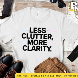 Puede incluir: Camiseta blanca con el texto "LESS CLUTTER, MORE CLARITY" en negro. El texto está subrayado con líneas azules y rosas. La camiseta se muestra sobre una superficie de madera con accesorios.