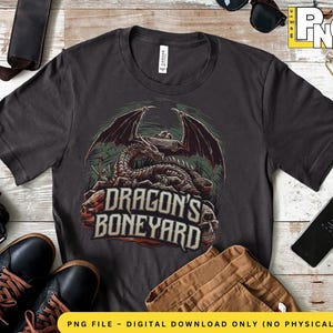 Peut inclure: Un t-shirt gris foncé avec un motif graphique d'un squelette de dragon avec des ailes et le texte "Dragon's Boneyard".