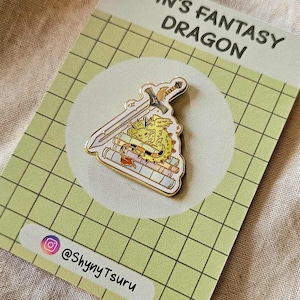 Peut inclure: Épingle en émail doré représentant un dragon, une épée et des livres. L'épingle est posée sur une carte à grille verte avec le texte "Pin's Fantasy Dragon" et le compte Instagram "@ShynyTsuru".