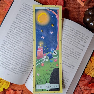 Peut inclure: Marque-page rectangulaire à bordure jaune avec une illustration fantaisiste. Le dessin comprend un croissant de lune, des étoiles, des papillons, des fleurs et un squelette. Le texte "THE READER" est en bas. Le marque-page est posé sur un livre ouvert.