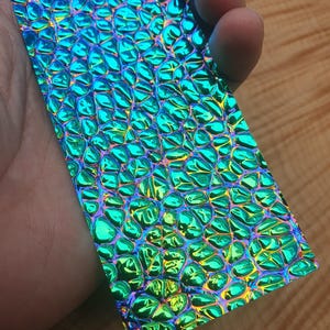 Puede incluir: Una lámina de vinilo holográfico verde y azul iridiscente con un patrón texturizado en relieve de piel de cocodrilo.