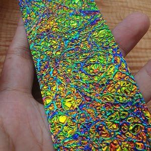 Op de afbeelding: Een rechthoekig stuk iriserend materiaal met een textuur, bubbelachtig oppervlak. De kleuren verschuiven van geel en groen naar blauw en paars, wat een levendig effect creëert. Het materiaal wordt in een hand gehouden.
