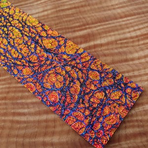 Pode incluir: Uma peça retangular com um padrão vibrante e intrincado de laranja, amarelo e azul. O design apresenta uma rede de formas interconectadas, que se assemelham a uma vista de perto de uma textura natural. A peça está sobre um fundo de madeira.