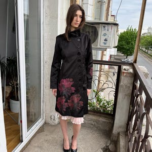 May include: A black knee-length coat featuring a high collar, a side-button closure, and dark red floral patterns on the lower front and sleeve. The coat is worn over a light pink pleated skirt and black pointed-toe heels, creating a layered look.