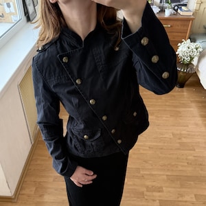 Puede incluir: Una chaqueta negra de estilo militar con cuello alto, mangas largas con tres botones en cada puño y cierre frontal diagonal de botones. La chaqueta incluye dos bolsillos delanteros con solapas abotonadas y charreteras con botones.