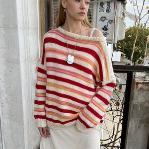 May include: A striped sweater in red, pink, orange, and white is worn with a white slip skirt and white sneakers with black stripes. A silver pendant hangs from a necklace. The sweater has a relaxed fit.